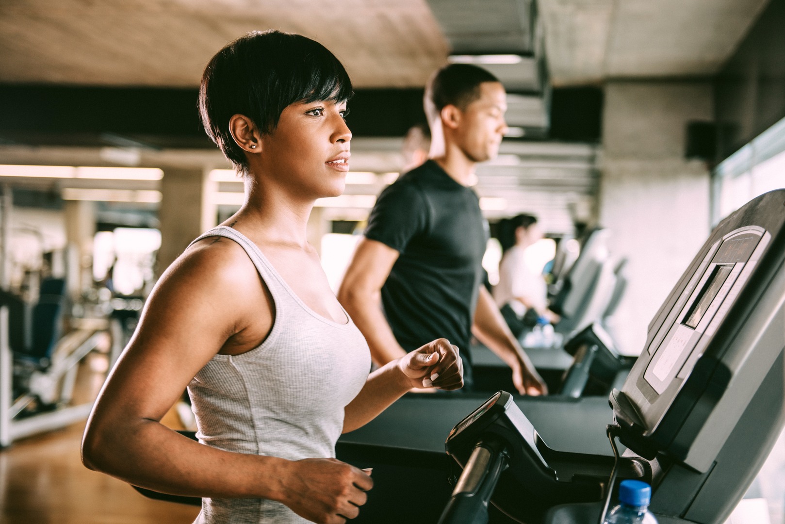 A Black woman running on a treadmill with other residents in a warmly lit fitness center complete with cardio, weight training and yoga room.